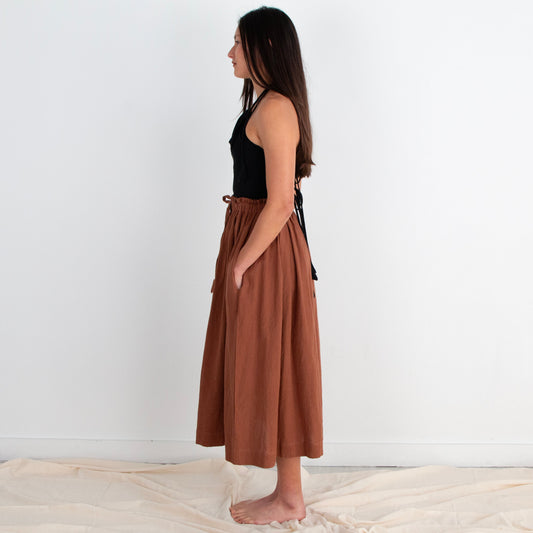 Woman wearing a black top and brown skirt standing against a white wall.