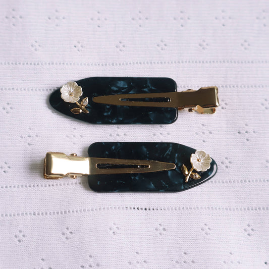 Two black hair clips with gold accents and floral decorations on a white textured background
