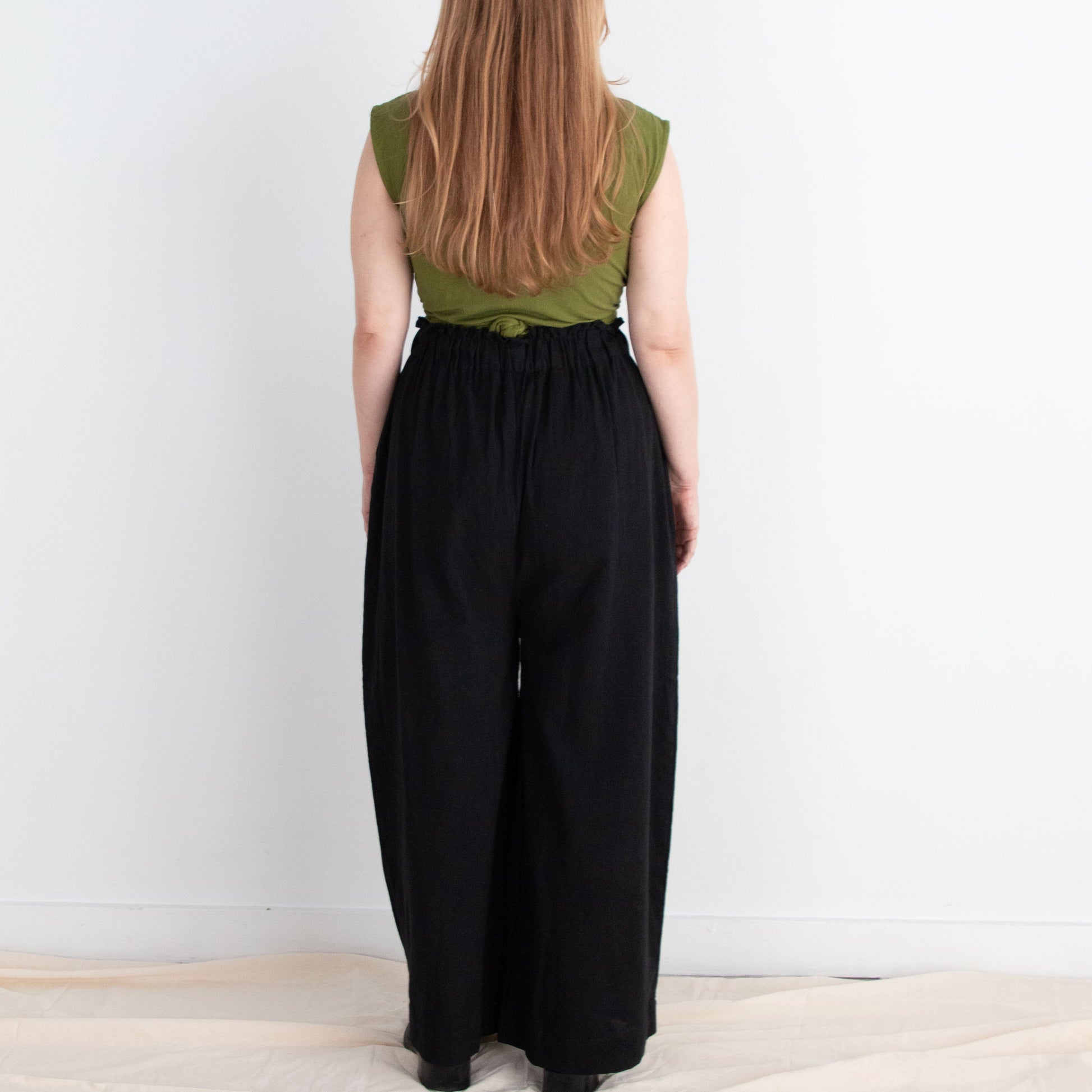Person wearing a green top and black pants standing against a white wall.