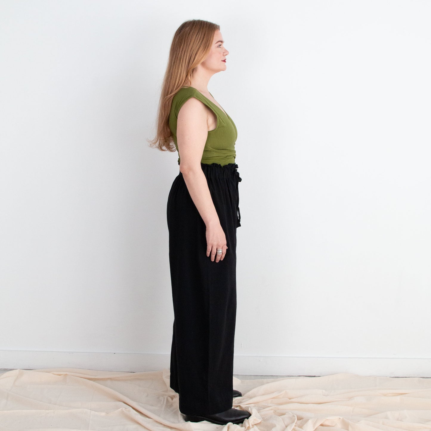 Woman wearing a green top and black pants standing against a white wall with draped fabric on the floor.