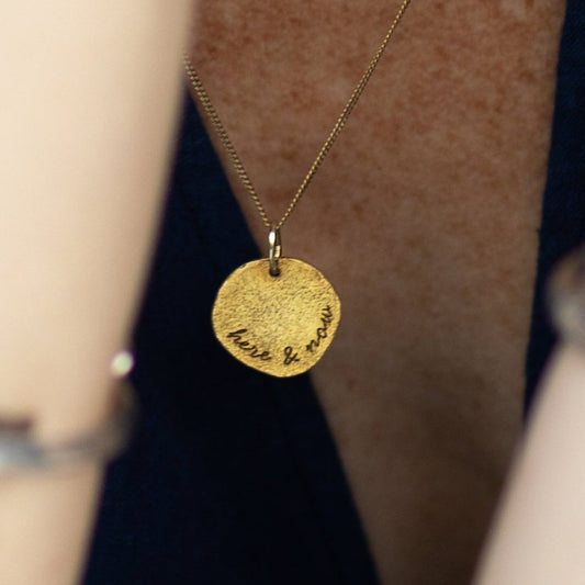 Gold necklace with a round disk, written "here & now", on a person wearing a dark top.