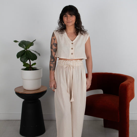 Woman in a white sleeveless top and pants standing next to a plant and chair against a white wall.
