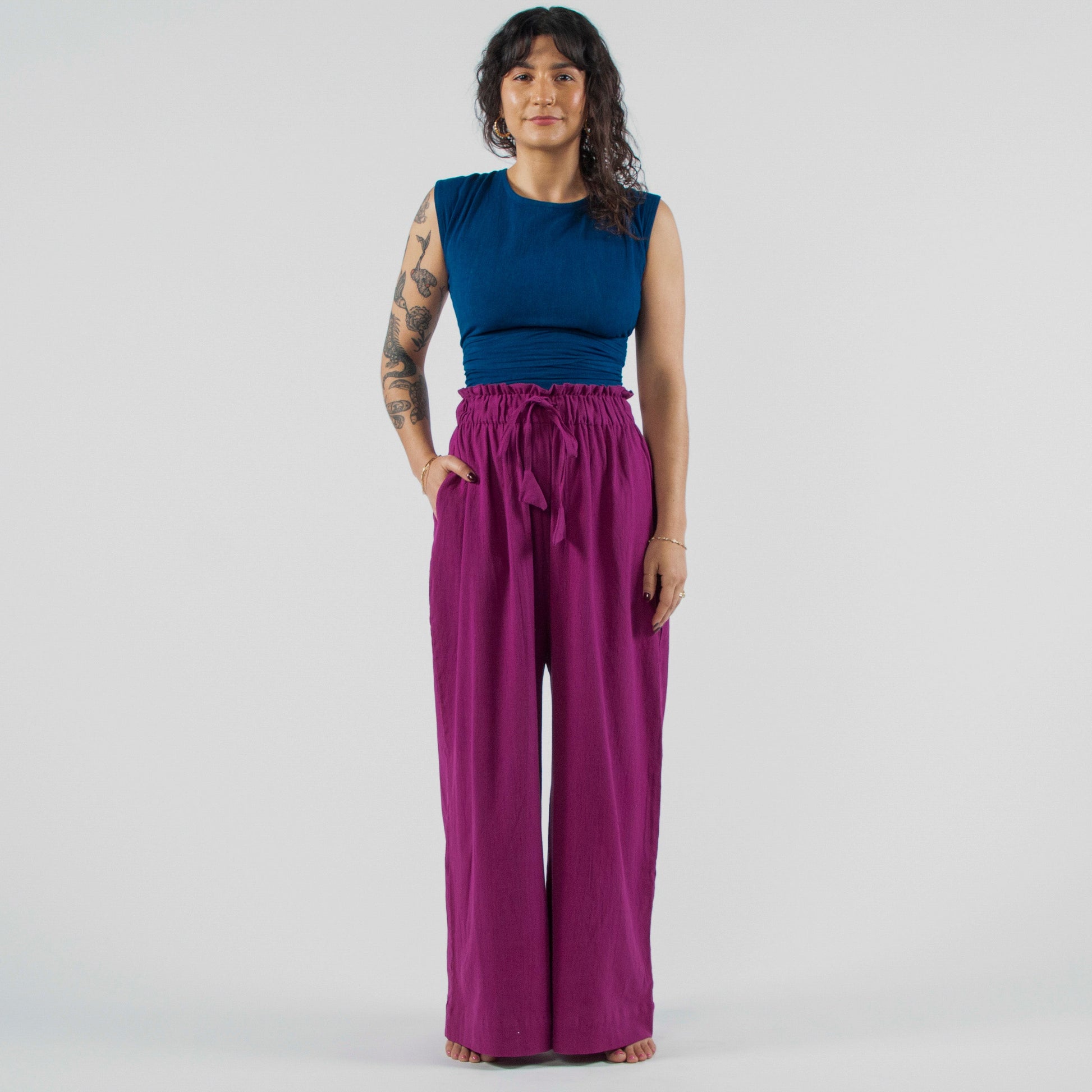 Woman wearing a blue sleeveless top and purple wide-leg pants standing on a white background.