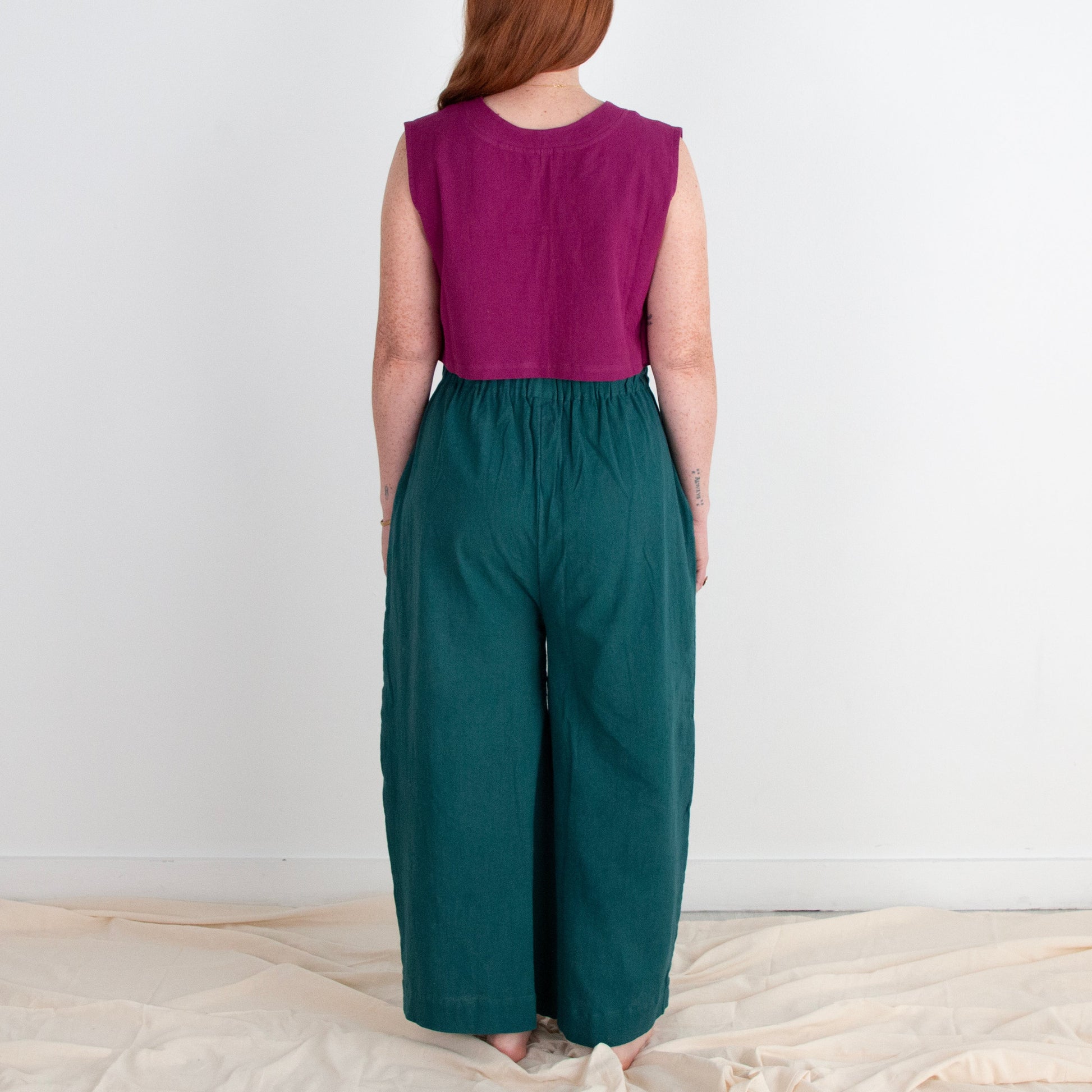Woman wearing a purple top and teal pants standing against a white wall.