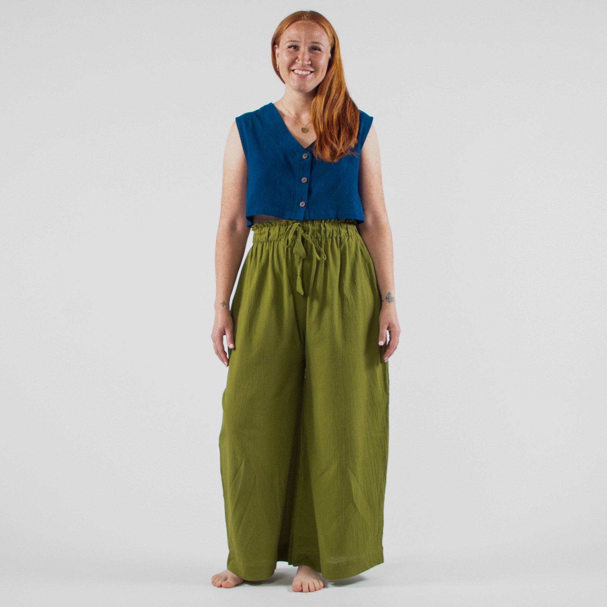 Woman wearing a blue sleeveless top and green wide-leg pants standing in front of a white backdrop with photography equipment.