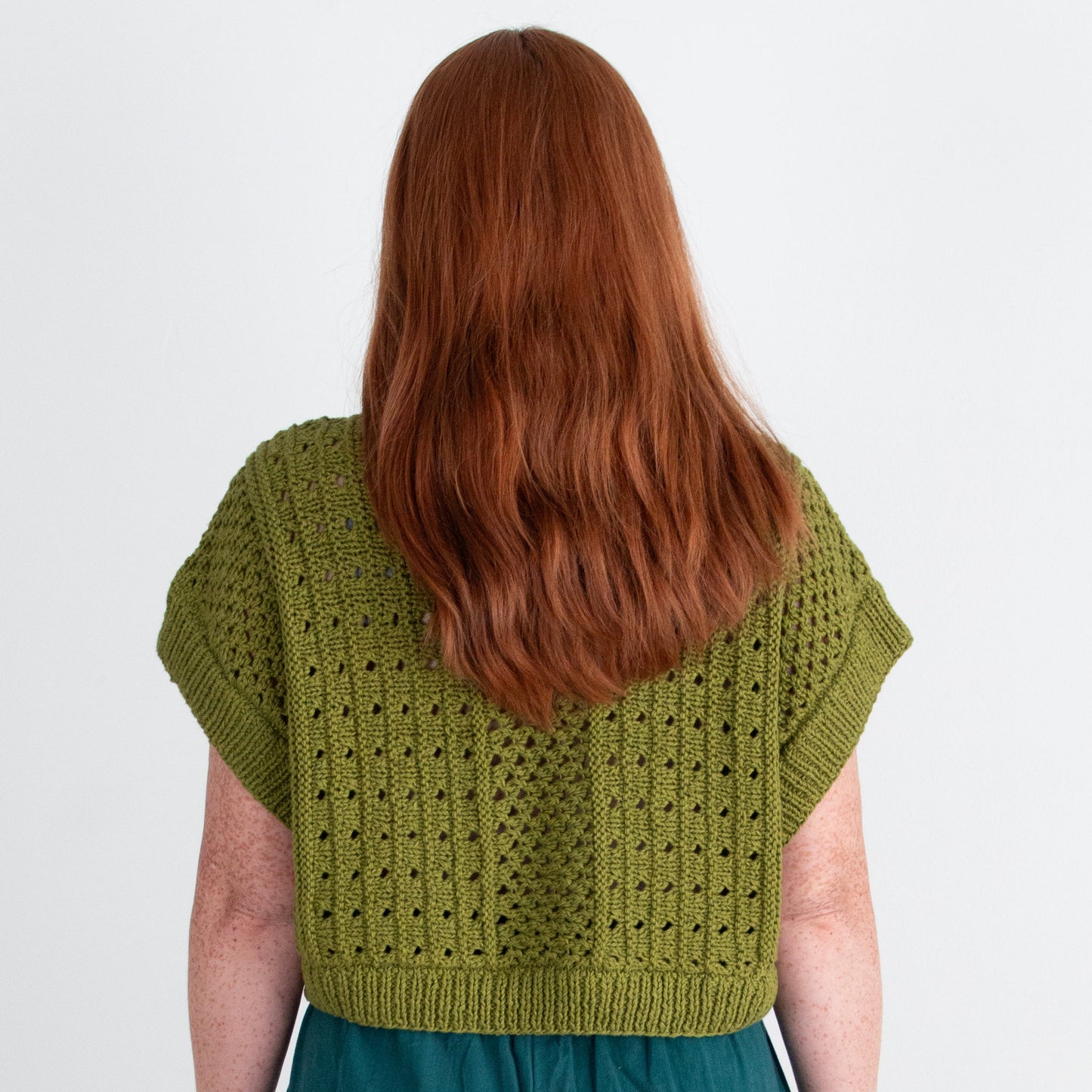 Person wearing a green knitted top and teal pants on a plain background
