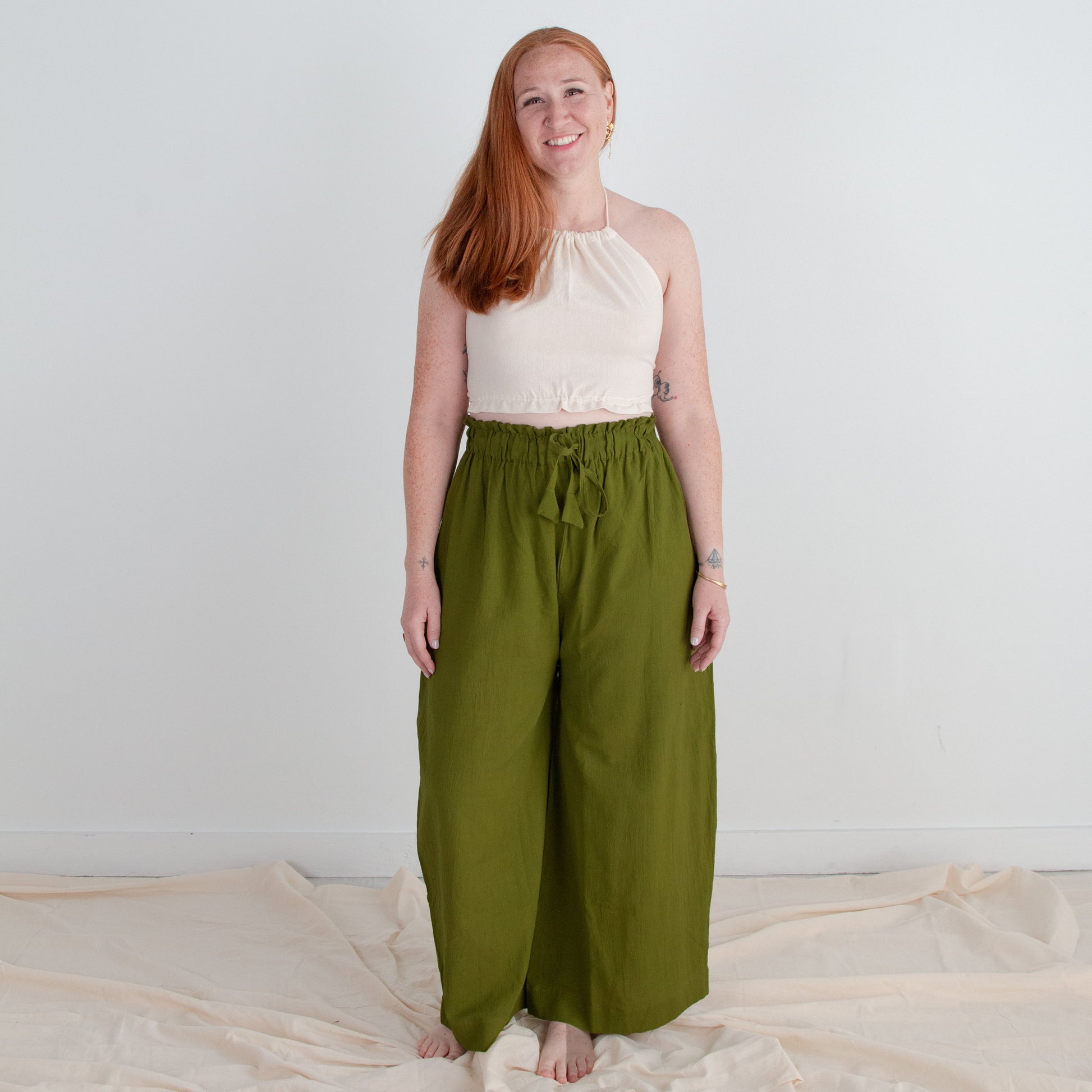 Woman wearing a white top and green pants standing on a light-colored fabric against a plain background